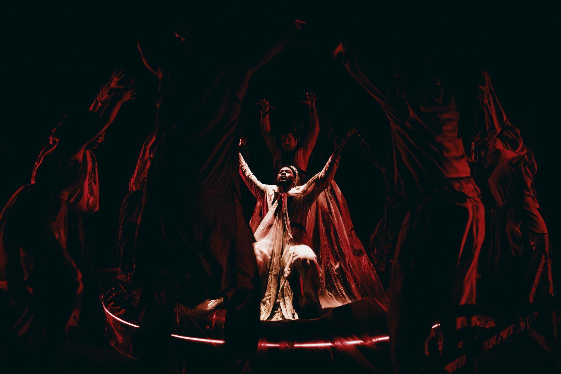 Performer in flowing white robes raises his arms centre stage, lit red, surrounded by silhouetted figures with arms upraised